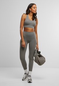 Grijze sportbh en bijpassende high-waisted legging, gemaakt van soepel, rekbaar materiaal. Model houdt een grijze gewatteerde tas vast, draagt witte sokken en sneakers.