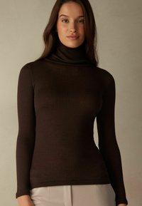 Hnědý žebrovaný svetr s dlouhým rukávem a rolákem, s přiléhavým střihem. Měkká textura a jemný lesk, v kombinaci s světlými kalhotami.