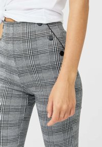 I pantaloni a quadri grigi a vita alta presentano dettagli in bottone neri lungo il lato, con una forma aderente e una texture di tessuto liscia.
