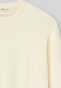 Pull oversized crème avec un col rond côtelé. Fabriqué en tissu tricoté doux, affichant une texture subtile et une coupe décontractée. Étiquettes visibles.