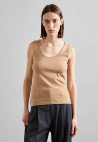 Pull sans manches beige en tissu doux et texturé avec un col en rond, porté avec un pantalon noir plissé. Design simple sans motifs.