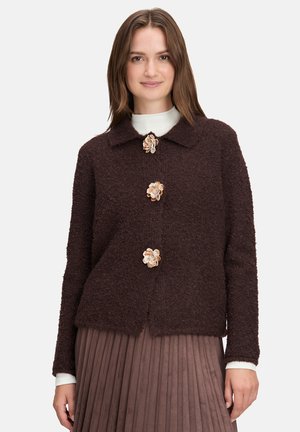 Braune strukturierte Jacke mit einem Kragen und drei dekorativen Blumenknöpfen auf der Vorderseite. Getragen über einem weißen Oberteil und einem plissierten Rock.