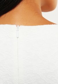 Weißer strukturierter Stoff mit einem subtilen Muster, ausgestattet mit einem kurzen Reißverschluss am oberen Rücken. Glattes und figurnahes Design.