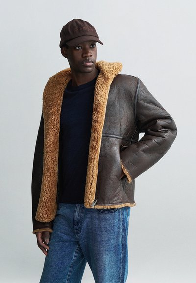 Veste en cuir marron avec un col et des finitions en shearling beige. Portée sur une chemise sombre et associée à un jean bleu. La casquette comporte des broderies.