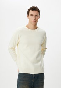 Cremfarbener Strickpullover mit rundem Ausschnitt und geripptem Saum, präsentiert mit gerade geschnittenen Jeans, vor einem schlichten Hintergrund.