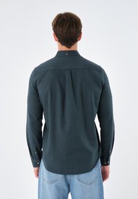 Uomo di spalle che indossa una camicia button-up a maniche lunghe di colore verde scuro e jeans in denim blu chiaro, su uno sfondo bianco semplice.