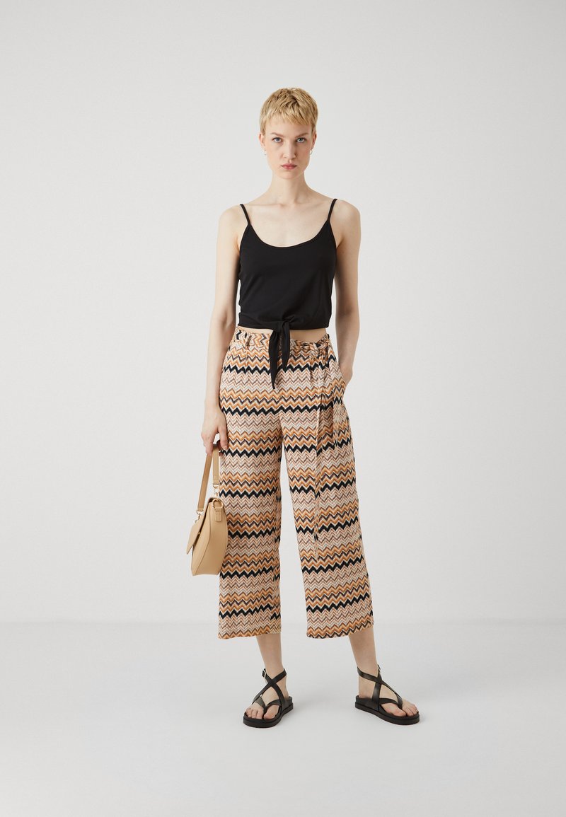 Čierny crop top s viazaním vpredu, skombinovaný so širokými nohavicami so vzorom v oranžovej, krémovej a čiernej zigzag dizajne. Obsahuje krémovú kabelku a čierne sandále.