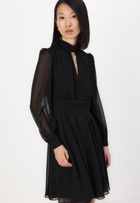 Fekete ruha magas nyakkivágással és kulcslyuk szabással, hosszú áttetsző ujjak, testhez simuló derék és fodros szoknya. Sima textúra és elegáns design.