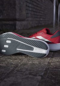 Zapatillas deportivas rojas con un upper texturizado, acentos grises y un logo blanco. La suela exterior presenta un patrón de tracción gris.