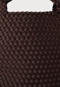 Καφέ υφαντή τσάντα με υφή στην επιφάνεια και ανθεκτικές λαβές, που διαθέτει πυκνό, περίπλοκο μοτίβο πλεγμένου υλικού.