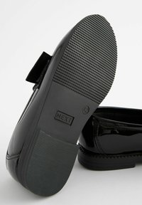 Chaussure noire en cuir verni avec une semelle en caoutchouc texturée, comportant un capuchon de chaussure lisse, des détails cousus et un petit nœud décoratif.