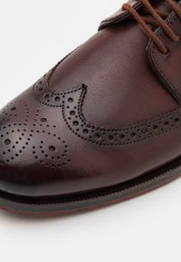 Chaussure habillée en cuir marron avec des détails de brogue, des perforations intriquées, une texture lisse et un bout arrondi. Design classique à lacets.
