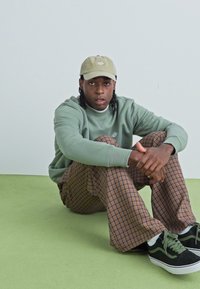 Grön sweatshirt, bruna rutiga vida byxor, svarta sneakers med gröna accenter och en beige keps. Sitter på ett ljust grönt golv.