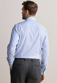Cămașă cu dungi albastre deschise, cu mânecă lungă, guler cu nasturi și butoni la manșete, purtată cu pantaloni gri. Textura fină din bumbac.