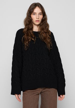 Žena s dugom valovitom smeđom kosom, odjevena u crni pulover s kabelskim pletenjem i smeđe hlače, na jednostavnoj sivoj pozadini.