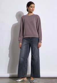 Felpa grigia a girocollo con dettagli di borchie, abbinata a jeans a gamba larga di colore scuro e scarpe flat argentate. Texture morbida e design casual.