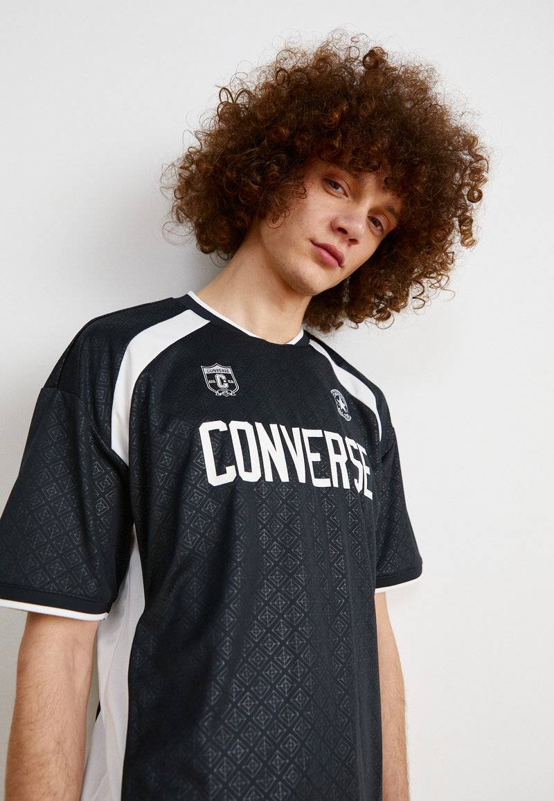 Camiseta deportiva negra con acentos blancos, que presenta un patrón de diamante texturizado y el texto "CONVERSE". Incluye un logo en el pecho.
