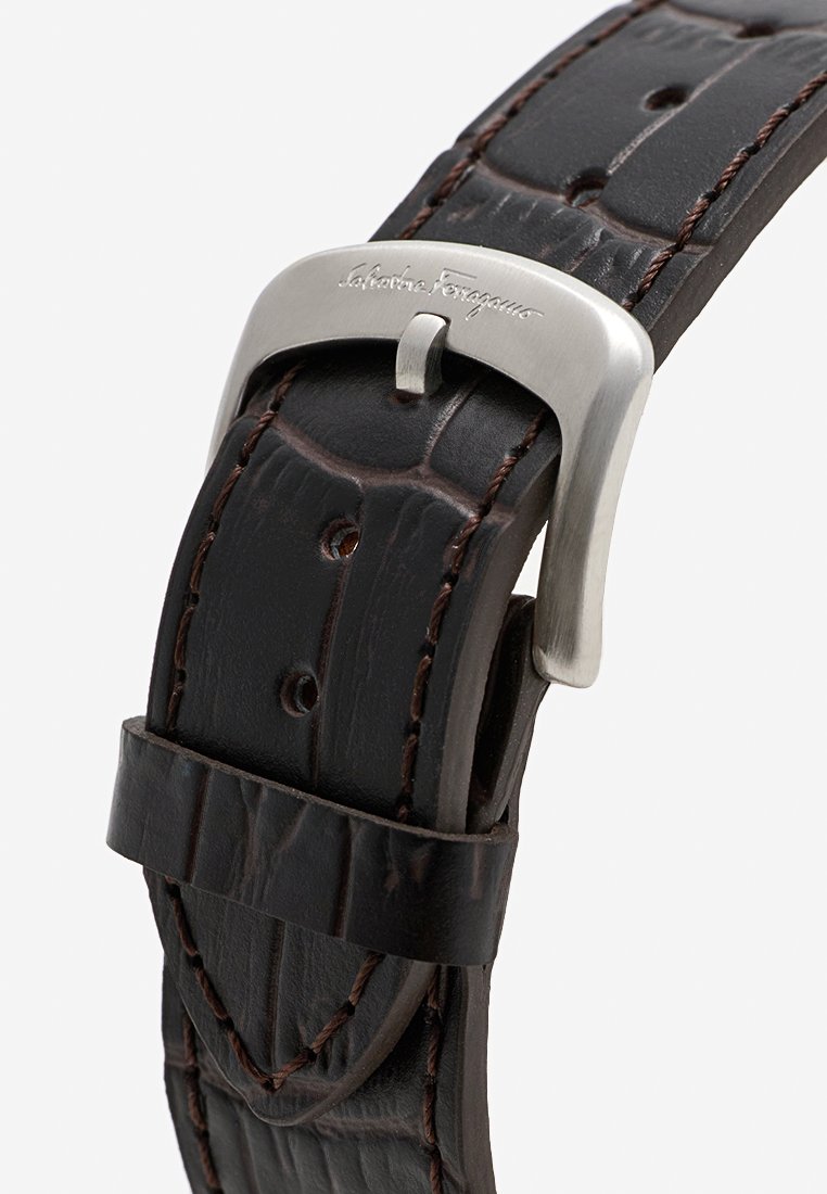 Bracelet de montre en cuir marron foncé avec texture crocodile et coutures, doté d'une boucle en métal argenté gravée du nom d'une marque de designer.