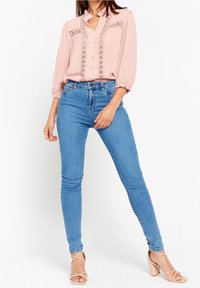 Kvinna i en blus med ljusrosa spetsdetaljer, hög midja blå smala jeans och nude blockklacksandaler, stående mot en vit bakgrund.