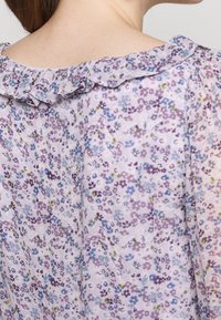 Blouse violet clair avec motif floral en bleu, vert et violet foncé. Comprend un col à volants et une texture de tissu fluide et ample.