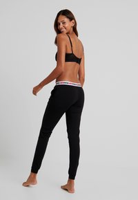 Schwarze Netz-Bralette mit verstellbaren Trägern, kombiniert mit schwarzen Jogginghosen, die einen schwarz-rot-gemusterten Bund tragen. Füße sind unbedeckt.