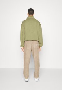 Olivgrön, krage sweatshirt med avslappnad passform; beige kordflöjelbyxor med två bakfickor; modell som står vänd bort, iförd vita sneakers.