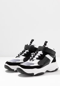 Masivní vysoké tenisky v černé, bílé a šedé barevné kombinaci, s síťovými panely, texturovanými akcenty a silnou gumovou podrážkou.