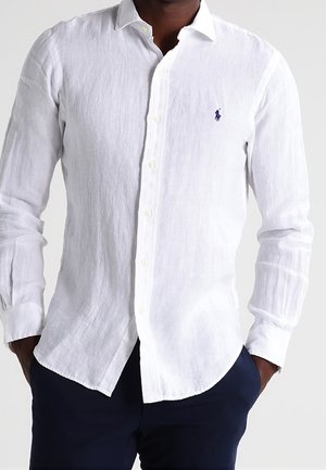 Man wearing a white long-sleeve button-up shirt with a small navy polo player logo on the left chest, paired with dark pants.