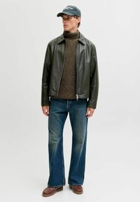 Giovane uomo che indossa una giacca di pelle verde scuro, un maglione a collo alto marrone, jeans blu, scarpe marroni e un cappellino nero mentre guarda di lato.