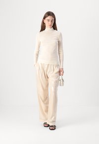 Maglietta lungo beige chiaro a maniche lunghe, colletto arricciato, abbinata a pantaloni larghi con paillettes dello stesso colore. Il modello tiene una piccola borsa bianca.