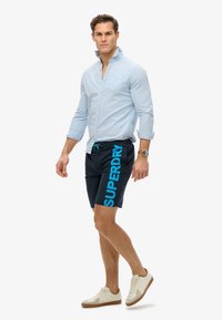 Camicia blu chiaro a maniche lunghe, pantaloni corti blu navy con "SUPERDRY" in blu brillante, e sneakers crema con suole marroni. Stile estivo casual.