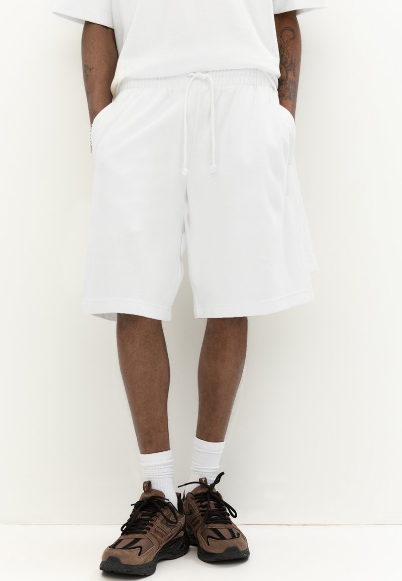 Shorts de sport blancs avec une taille élastique et un cordon de serrage, en tissu texturé. Associés à des baskets marron et des chaussettes blanches.