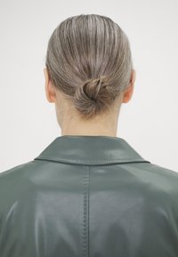 Personne aux cheveux gris vue de dos avec les cheveux en petit chignon portant une veste en cuir verte avec un col cousu.