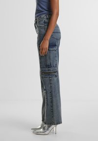 High-waisted blue denim cargo pants with multiple pockets, a loose fit, and visible seam detailing, paired with metallic heeled shoes.