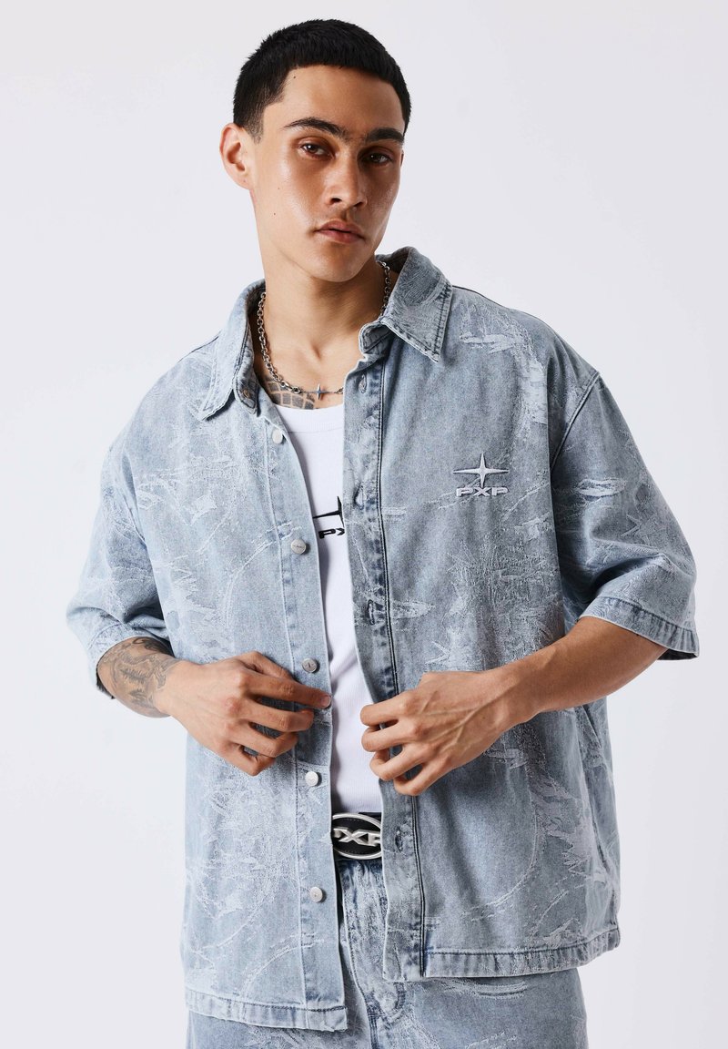 Jeune homme ajustant une veste en jean claire à manches courtes sur un t-shirt blanc, montrant son avant-bras tatoué et un collier en chaîne.