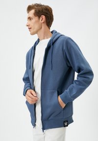 Sudadera azul con cremallera, bolsillo tipo canguro, capucha con cordón, y puños y dobladillo acanalados. Fabricada con un material suave y con una etiqueta de logo en la parte inferior.