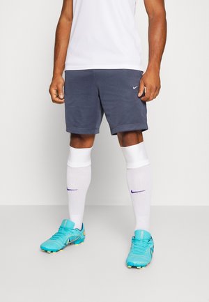 Homme portant des chaussettes de sport blanches, un short bleu foncé et des chaussures de football turquoise vif, debout devant un fond uni.