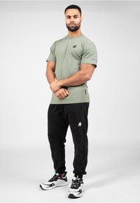 Olijfgroene katoenen T-shirt met een logo op de borst, gecombineerd met zwarte joggerbroeken met een wit logo en elastische boorden, en sportschoenen.