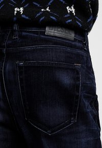 Jeans in denim blu scuro con una finitura testurizzata, caratterizzati da una patch rettangolare con il nome del marchio e tasche posteriori curvate.
