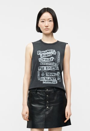 Jeune femme aux cheveux noirs et raides, portant un haut sans manches noir à motif graphique et une jupe en cuir noire à boutons sur le devant, debout devant un fond uni.