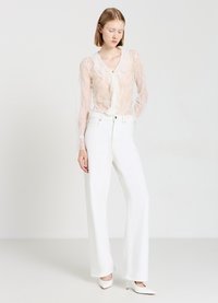 Blusa in pizzo color beige chiaro con chiusura a fiocco, abbinata a pantaloni bianchi a gamba larga. Le scarpe sono appuntite e bianche, creando un aspetto morbido e coeso.