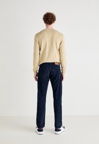 Man met krullend haar staat met zijn gezicht naar een witte muur, gekleed in een beige trui, donkerblauwe broek en witte sneakers met marineblauwe en rode details.