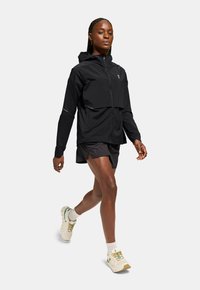 Lätt svart löpjacket med dragkedja framtill, huva och flera fickor; bärs med mörka shorts och neutrala atletiska skor.