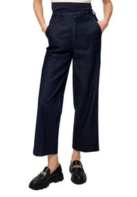 Person som bär mörkblå cropped vida byxor med framfickor, tillsammans med svarta loafers med guldkedjedetaljer.