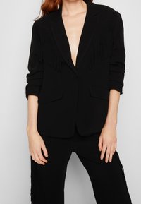 Černý blazer s třásněmi na ramenou, prošívanou strukturou, hlubokým výstřihem do V a dvěma předními kapsami. Dlouhé, vyhrnuté rukávy.