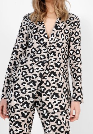 Donna che indossa un blazer beige e nero con stampa leopardata e pantaloni abbinati, con un bottone chiuso e mani rilassate lungo i fianchi.