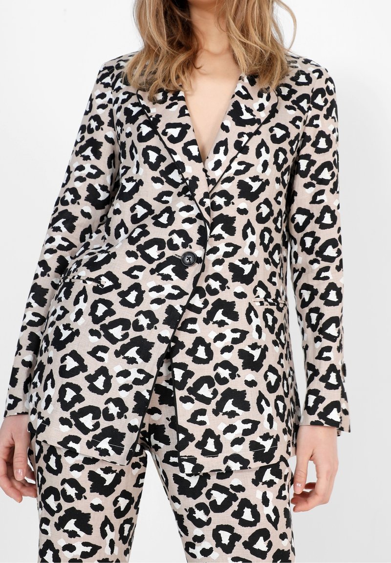 Femme portant un blazer beige et noir à imprimé léopard et un pantalon assorti, avec un bouton fermé et les mains détendues le long du corps.