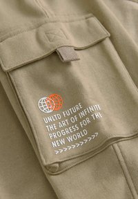 Khaki tyg med en slät textur. Har en ficka med en design av överlappande cirklar i vitt och orange, samt tryckt text i fet vitt.