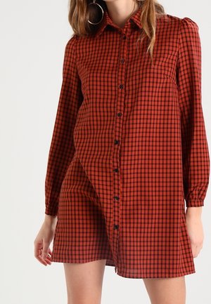 Robe-chemise à manches longues à carreaux rouges et noirs, boutonnée, portée par une personne debout, coupée au-dessus des genoux, avec de grandes boucles d'oreilles en créole.