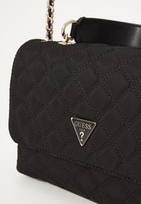 Svart quilted håndveske med et triangulært gulllogo, lenke-håndtak og glatte svarte skinnakenter. Den teksturerte overflaten har diamantmønstre.